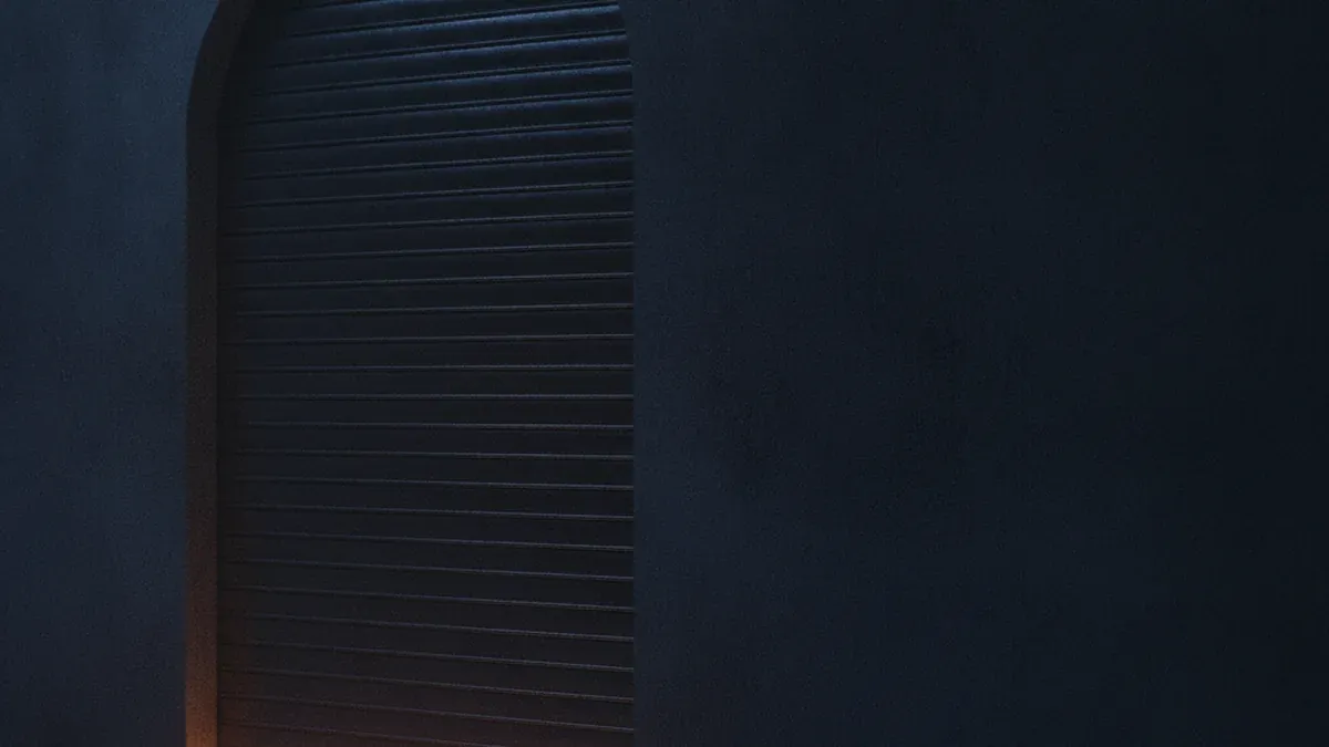 A heavy dark metal shutter closed fully across a doorway, warm light pressing through the narrow gap along the floor without getting in