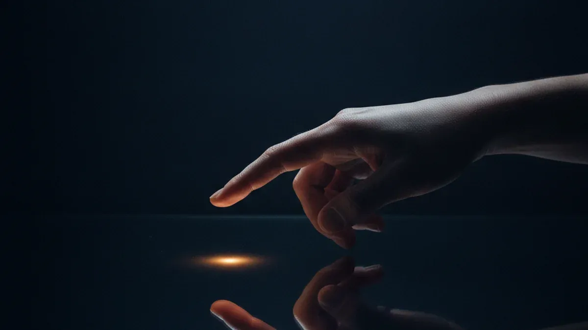 A single hand hovers with an extended fingertip a few centimetres above a dark blank glass surface which reflects the finger back as a soft mirrored shape