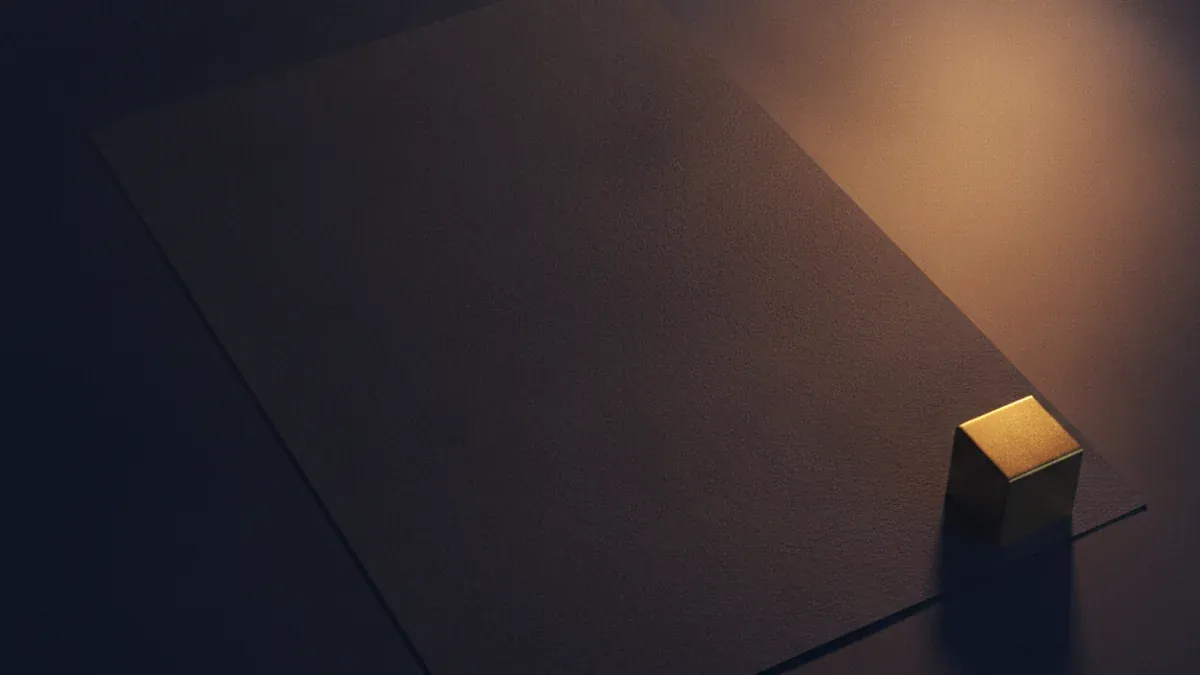 A single blank dark sheet of paper lying square on a desk in the warm beam of a lamp, weighted at one corner by a plain brass paperweight
